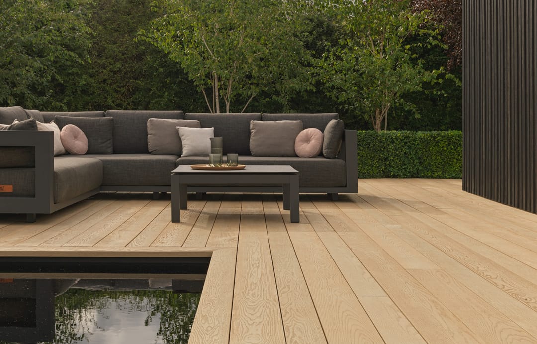 Pale Ashwood deck edging a reflecting pool, with a grey outdoor sectional sofa and trees behind a dark timber screen