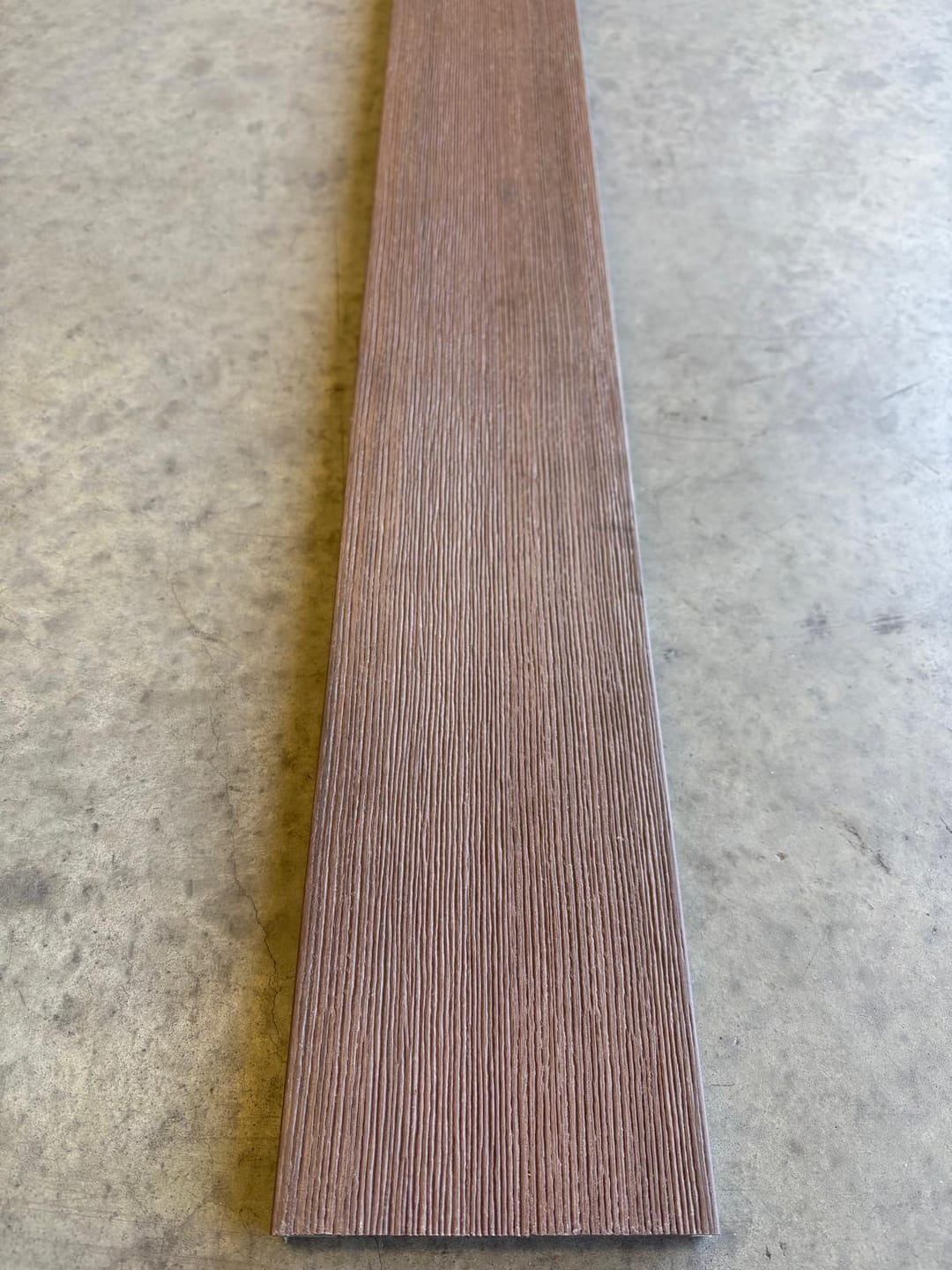 Modelo Prime Merbau solid composite deck board lying on a concrete floor, showing its red-brown brushed wood-grain face