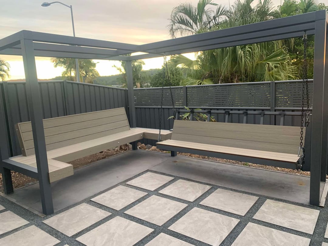 Built-in bench seats clad in pale blonde composite boards under a dark pergola on a paved courtyard