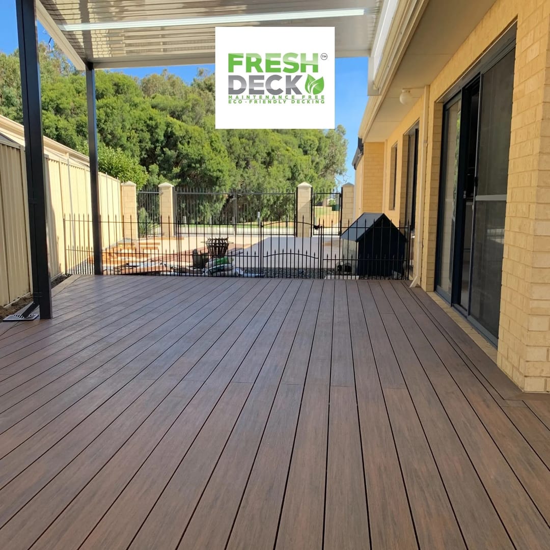 Brown FreshDeck composite patio deck under a pergola along a cream brick house, with a pool fence beyond