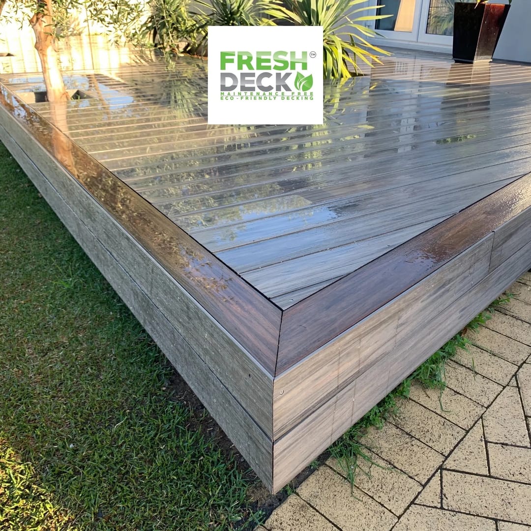 Rain-wet raised FreshDeck composite deck with a picture-frame border and fascia, set on a lawn beside brick paving