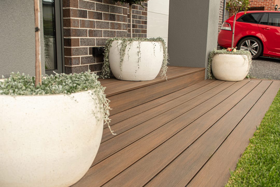 Warm brown WoodEvo composite deck at a brick home entry, with large white planters and a lawn edge