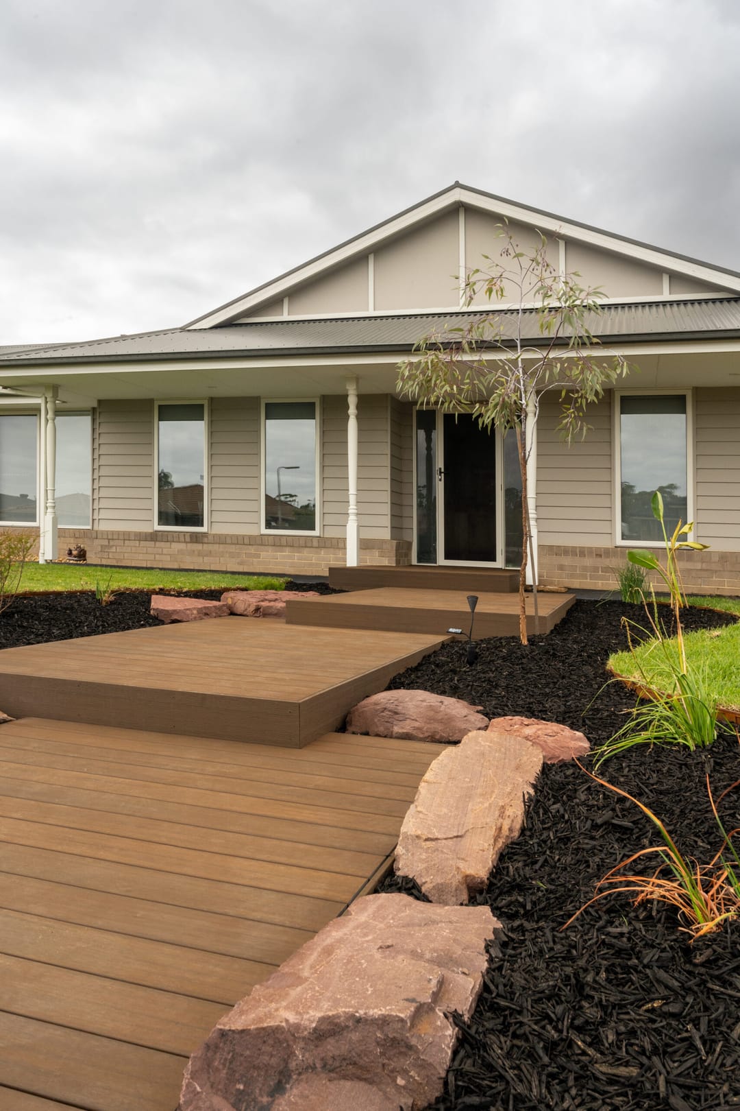 Brown WoodEvo HOME composite deck platforms leading to a farmhouse front door, edged with sandstone rocks and mulch
