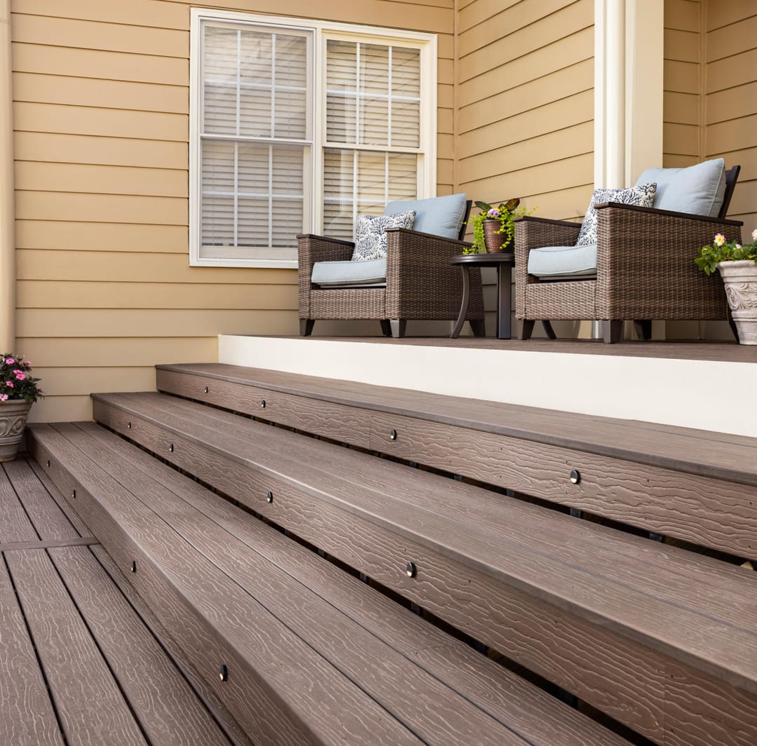 Wide Cold Brew steps rising to a raised deck with two wicker armchairs against a pale yellow weatherboard house
