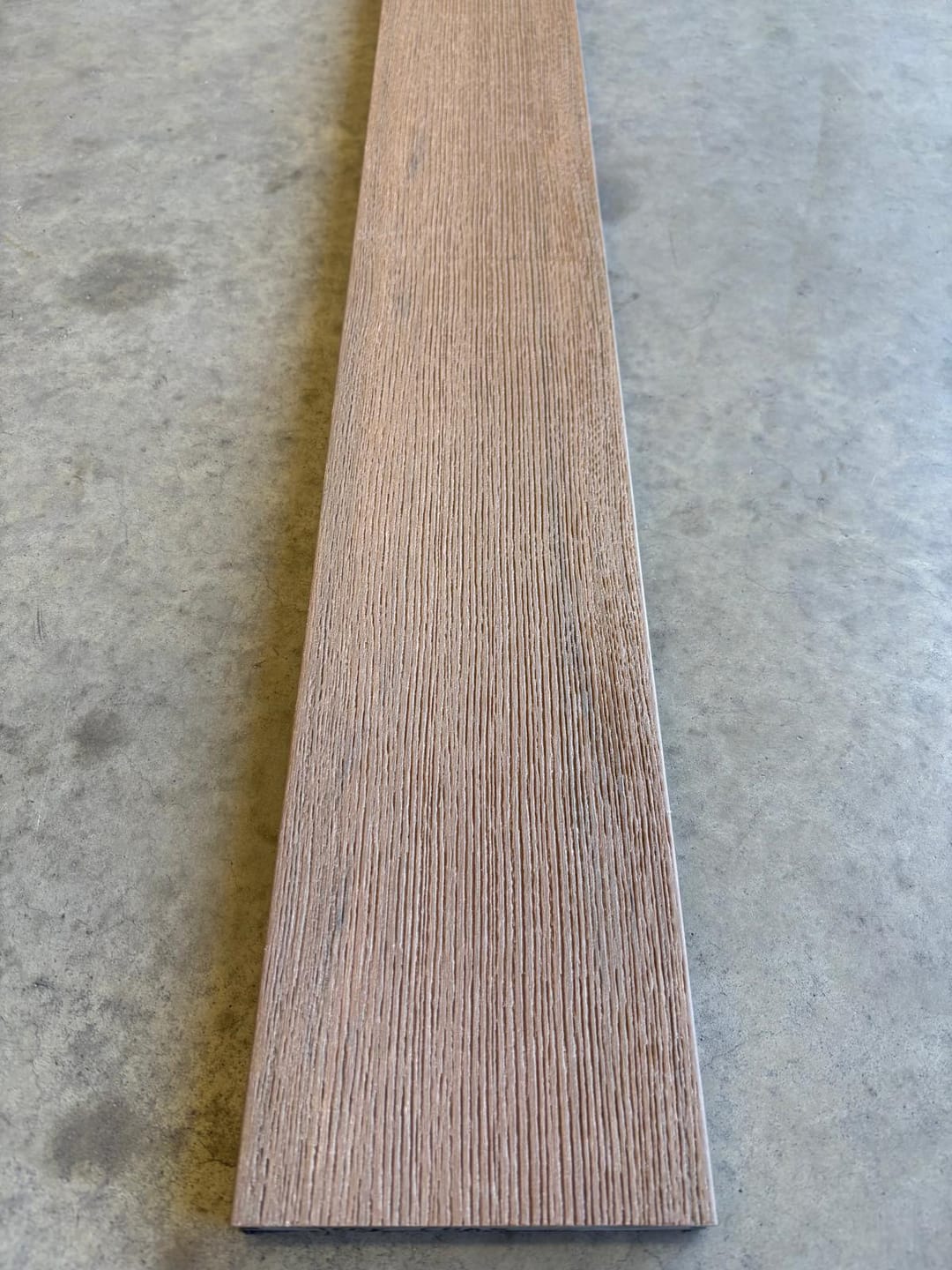 Modelo Prime Teak solid composite deck board lying on a concrete floor, showing its light pinkish-brown brushed wood-grain face