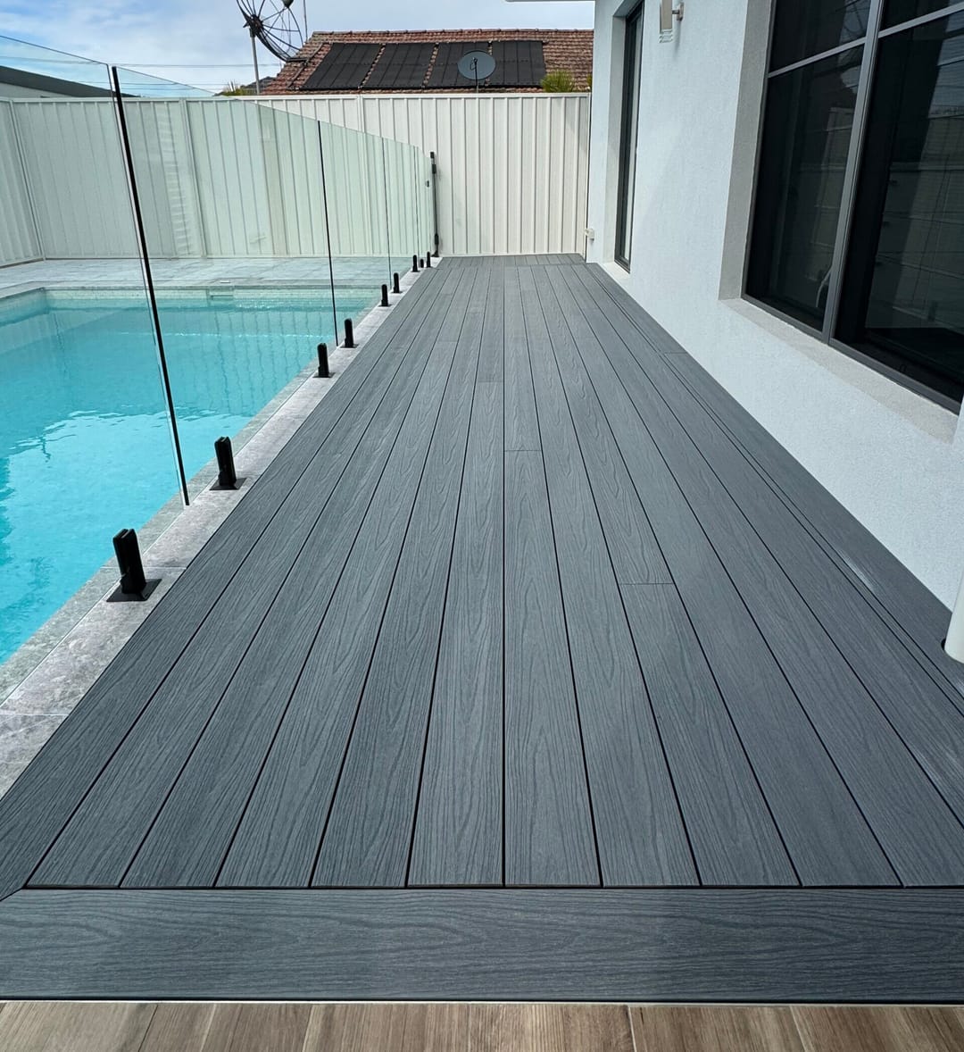 Long charcoal-grey Acetech deck walkway between a house and a pool with frameless glass fencing