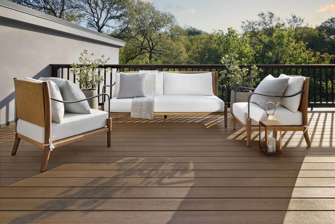 Brown English Walnut deck with a white outdoor sofa and armchairs behind a black railing, treetops beyond