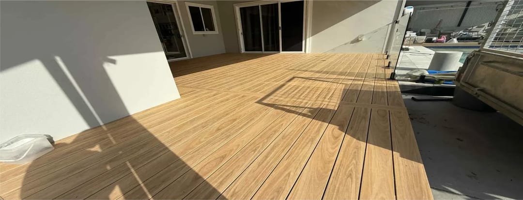 Newly laid honey-toned Origin composite deck under a house overhang with sliding doors and a pool fence