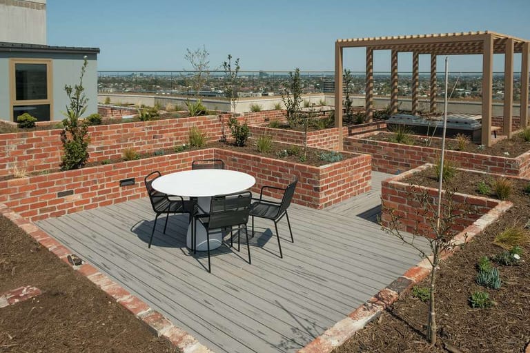 Grey ModWood Natural Grain Silver Gum deck with a round outdoor table, set among raised brick garden beds with a timber pergola