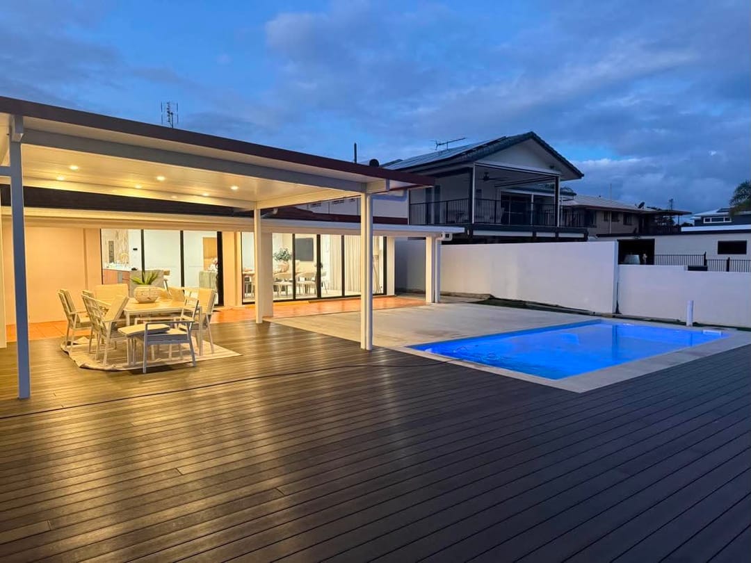 Dark brown composite pool deck at dusk wrapping a lit swimming pool and a covered alfresco dining area