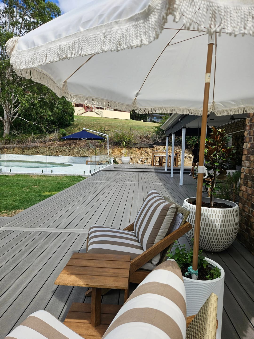 Grey composite deck with striped sun lounges and a fringed umbrella, a pool and lawn beyond