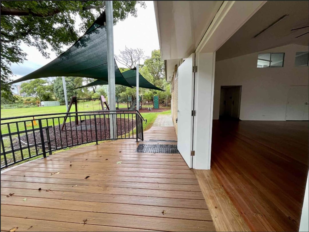 Blackbutt deck along a community hall with open doors, black railing and shade sails over a park