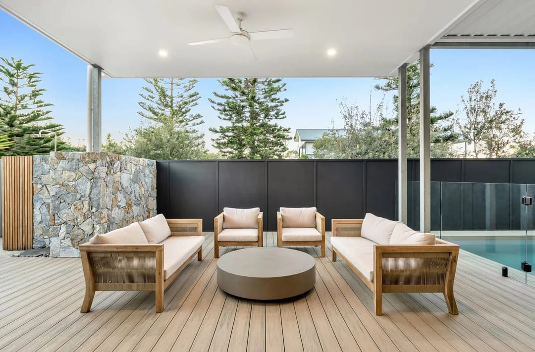 Covered alfresco deck in a pale warm-grey composite with outdoor lounges, a stone feature wall and a pool behind glass