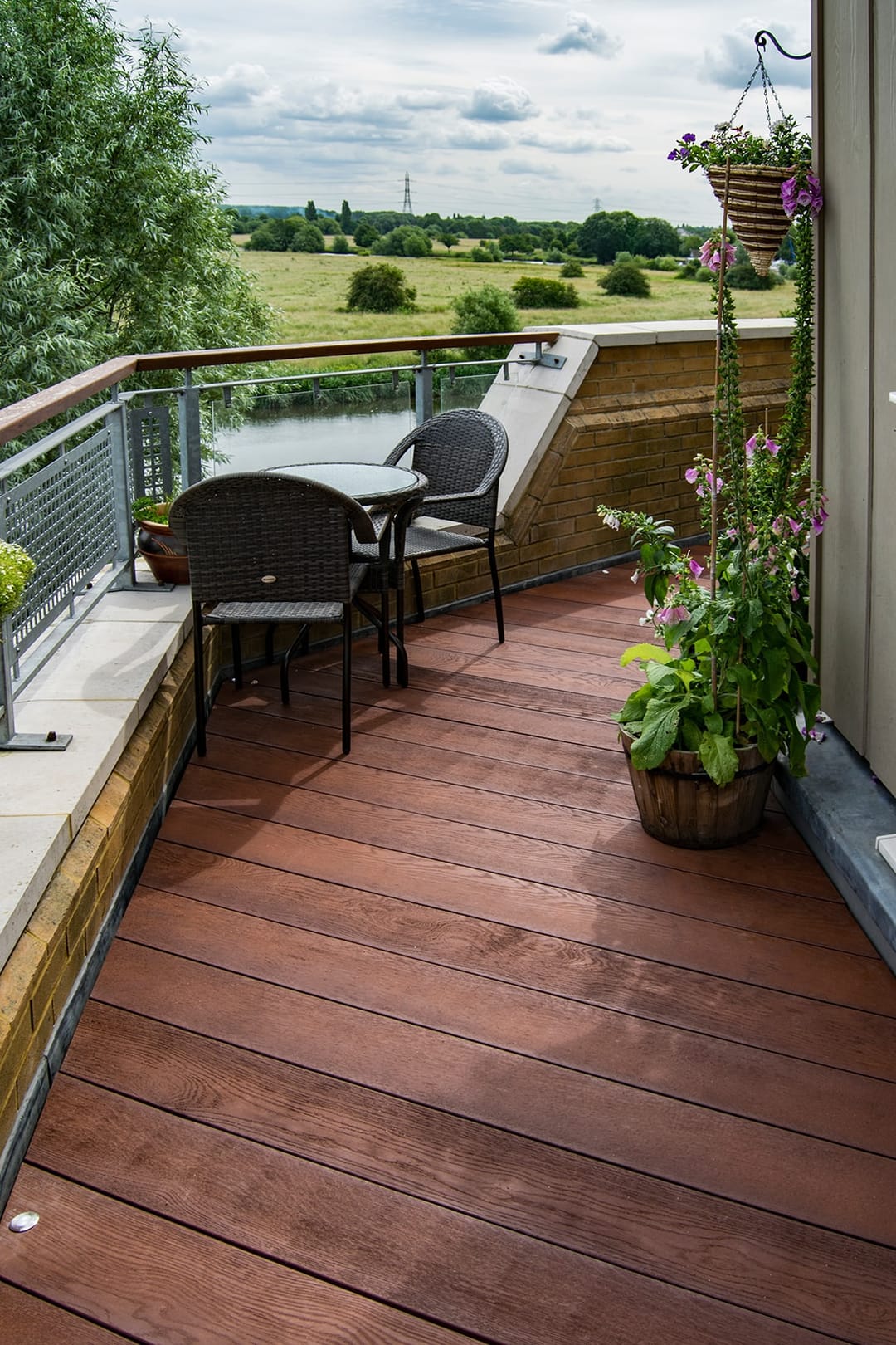Narrow balcony decked in red-brown Jarrah boards with a bistro set, hanging baskets and a view over parkland