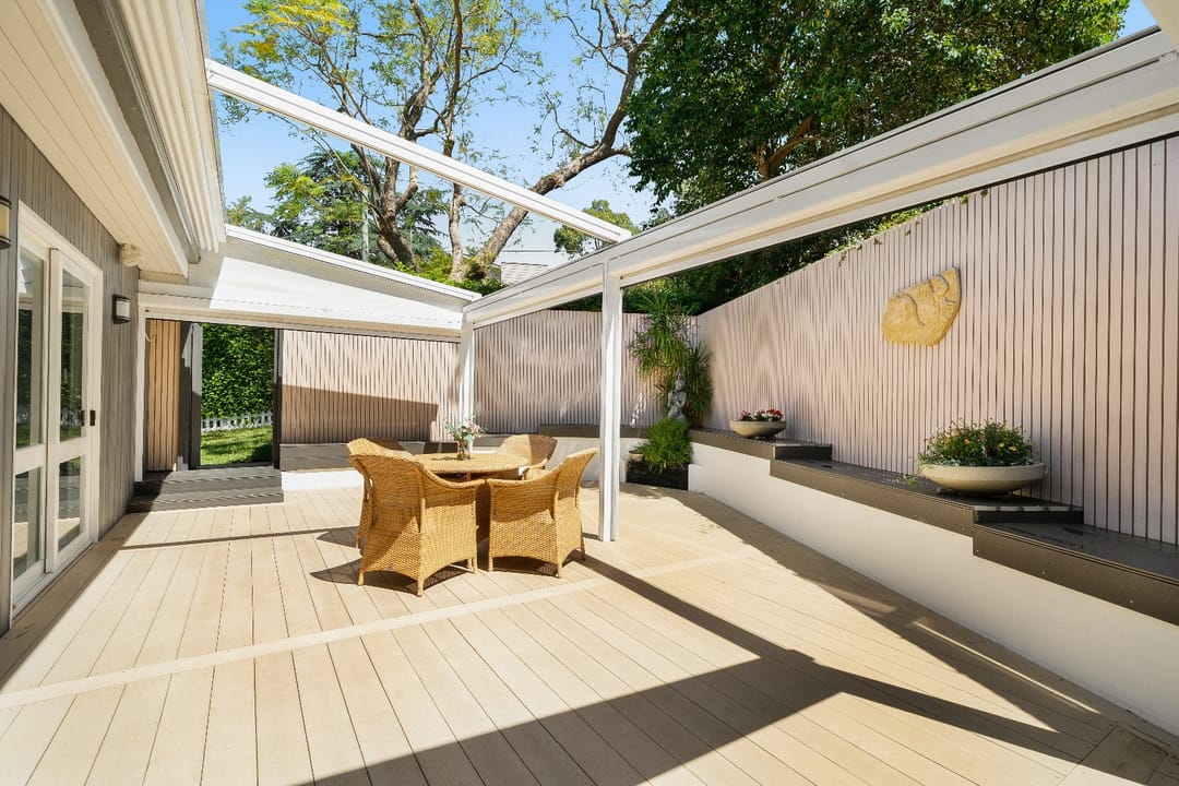 Pale INNODECK courtyard deck under an open white pergola, with a rattan dining setting, slatted screen walls and garden beyond
