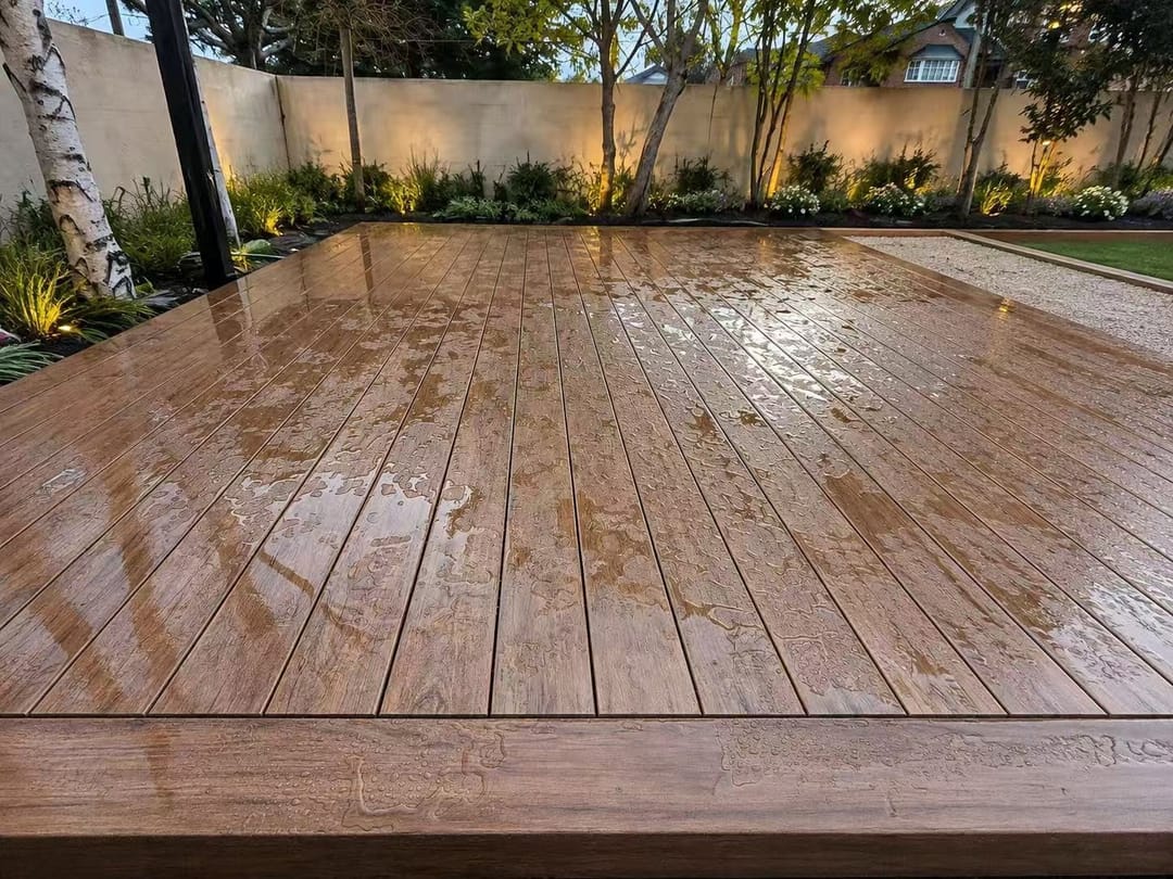 Rain-wet Evertex Vivid Classic Merbau deck with a picture-frame border, lit garden beds and birch trees behind