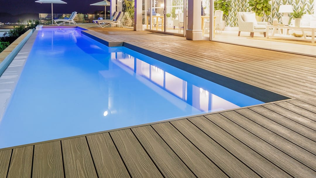 Long lit swimming pool at dusk edged with Eva-Last Infinity Oasis Palm decking beside a glass-walled home