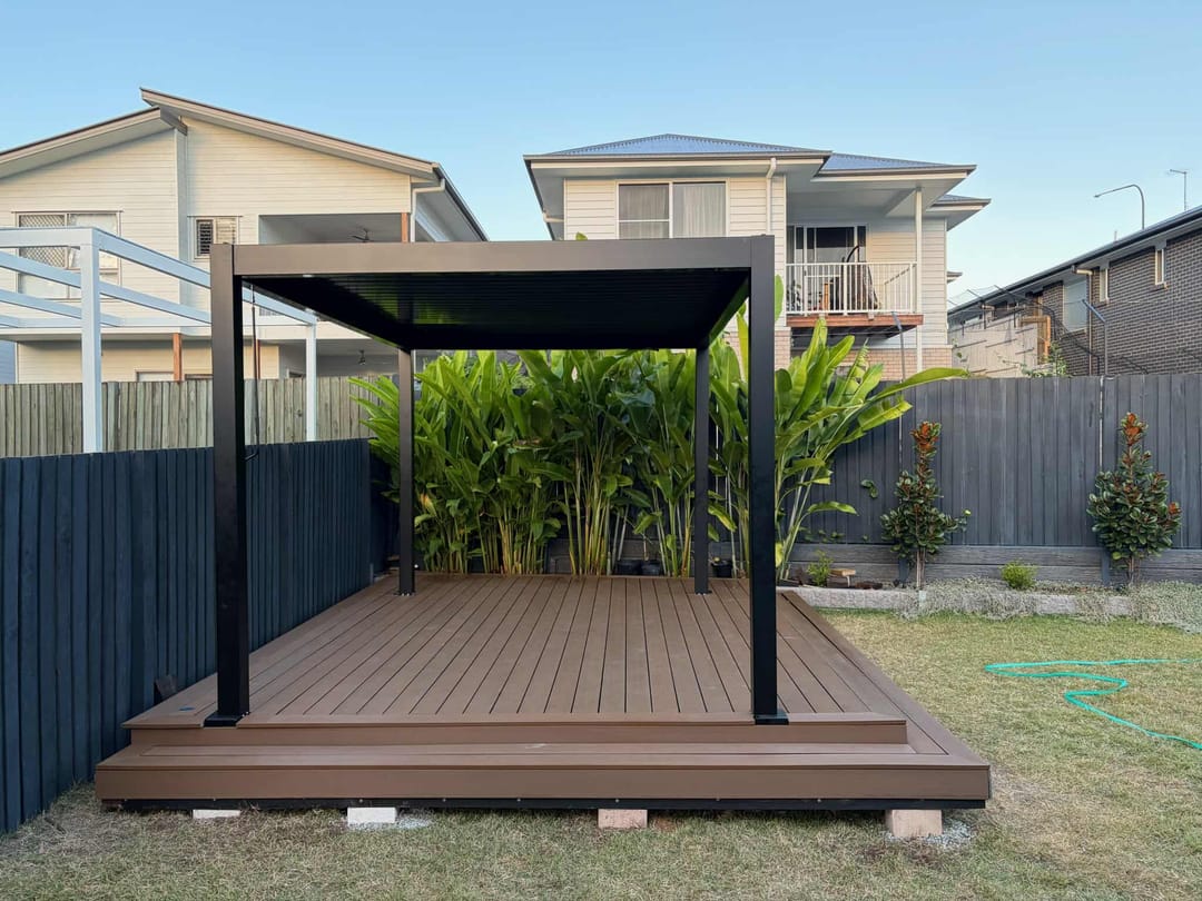 Reddish-brown composite deck platform under a black pergola with tropical plants in a suburban backyard