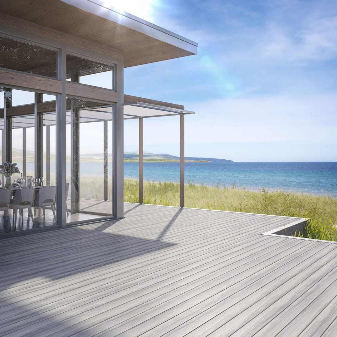 Pale grey Amalfi deck wrapping a glass-walled beach house, with dune grass and the sea beyond