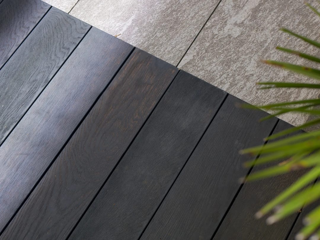 Close view of near-black Burnt Cedar decking meeting grey paving, with a palm frond in the corner