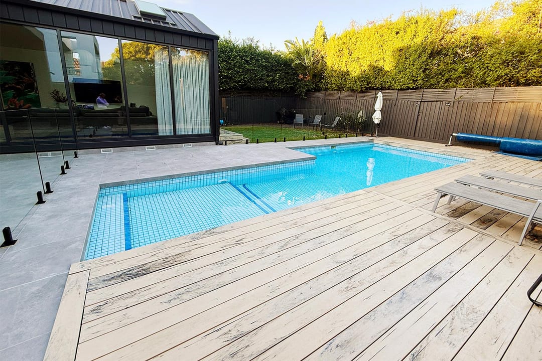Light grey OneDeck PVC deck wrapping a rectangular pool with glass fencing and a modern black-framed house