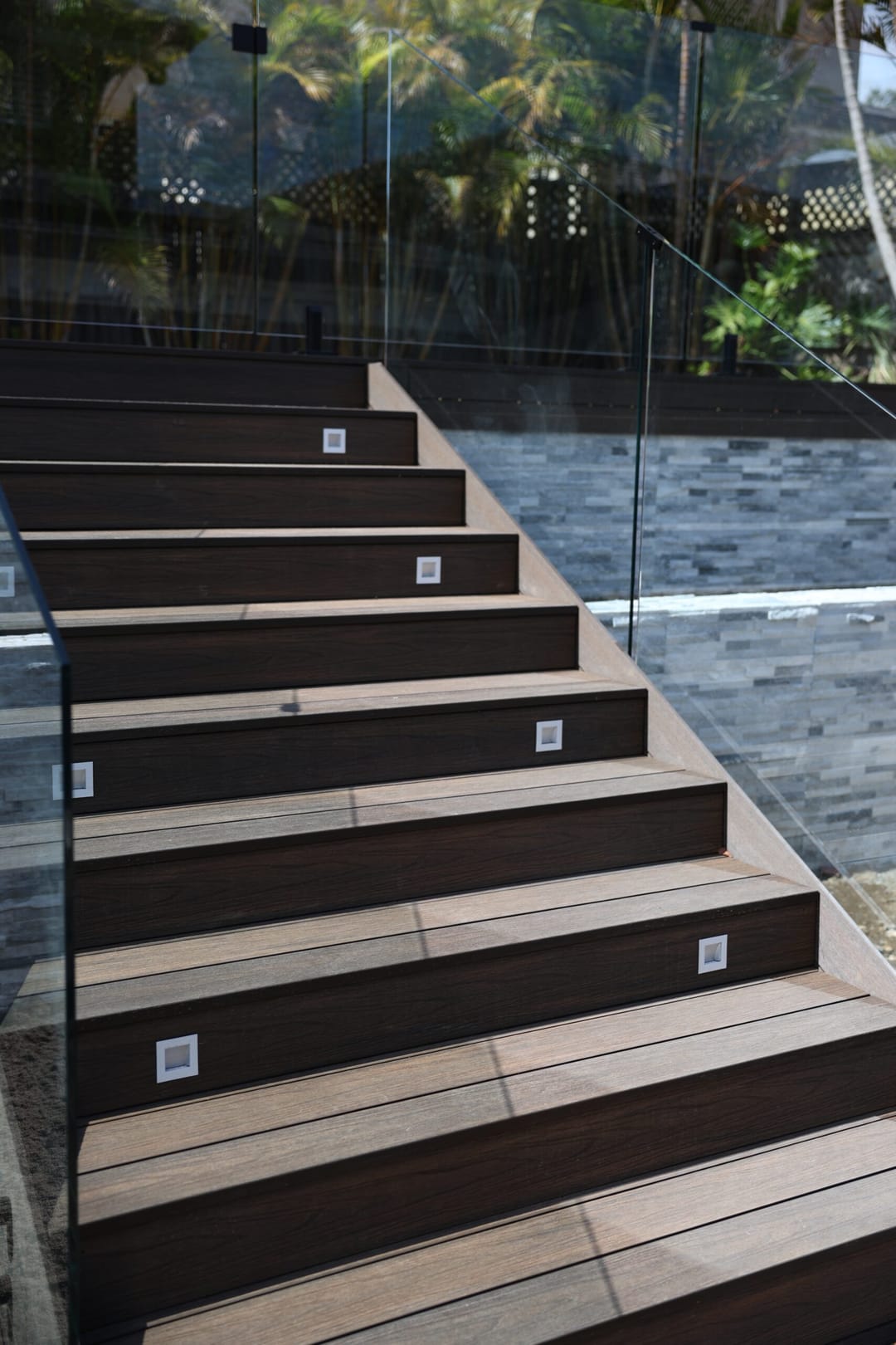 Dark brown Acetech composite stair treads with inset step lights, beside a stacked-stone wall and glass balustrade