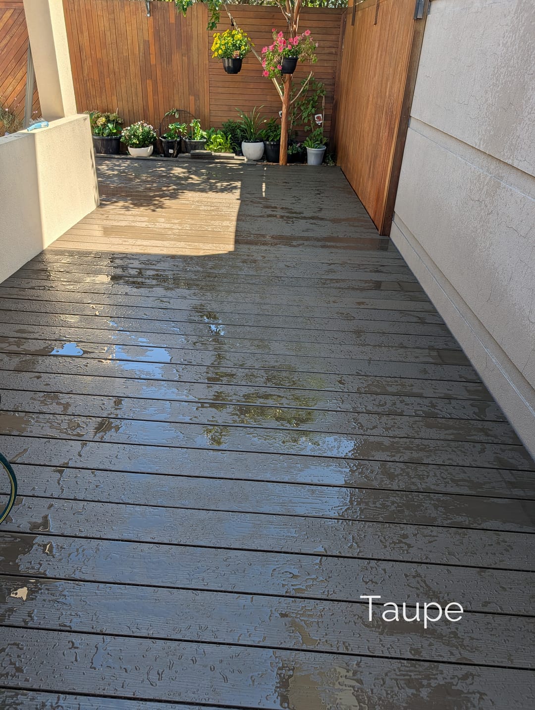Wet Evertex Essence Taupe deck in a narrow courtyard between a timber fence and a rendered wall, with potted plants
