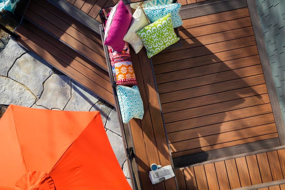 Overhead view of a reddish Mahogany deck with a cushioned bench, bright pillows and an orange umbrella beside stone paving