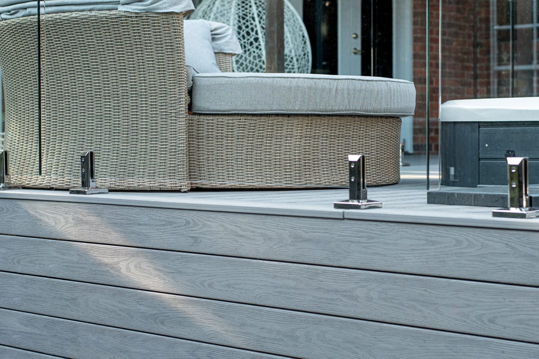 Light grey composite deck with glass pool-style fencing, a wicker daybed and a spa beside a brick home