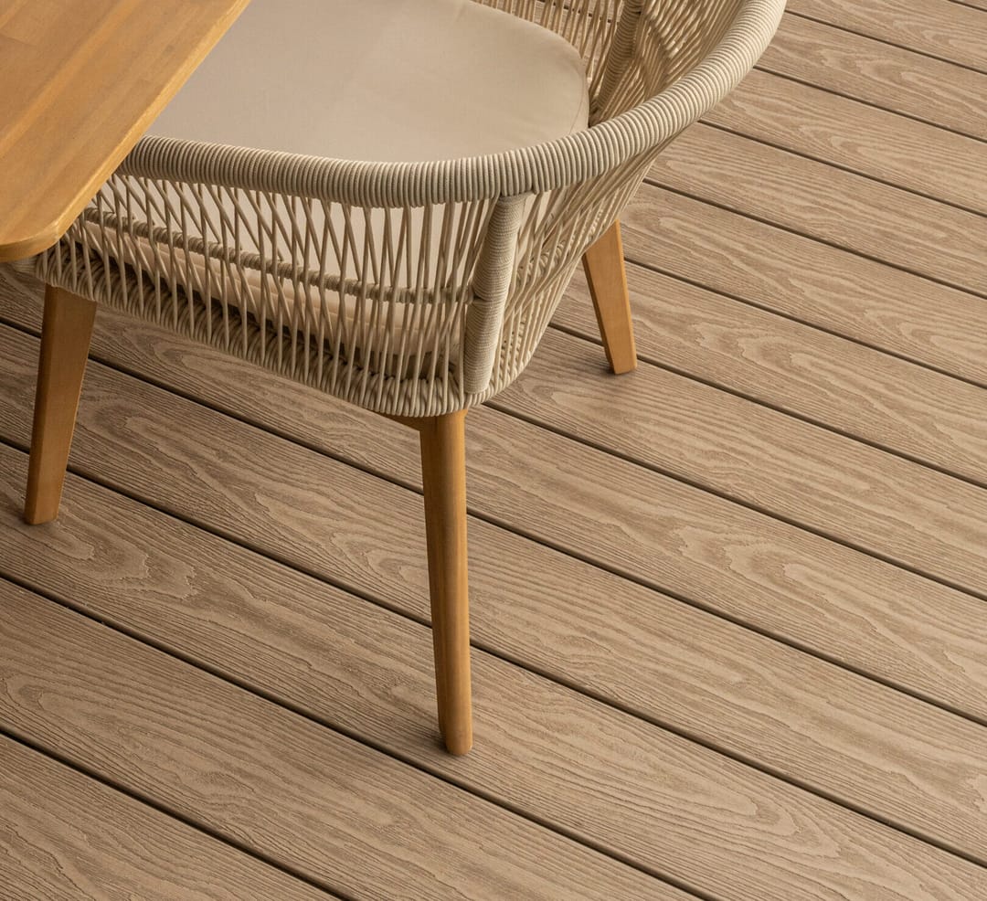 Close-up of pale sandy-beige composite decking boards under a woven outdoor dining chair and timber table