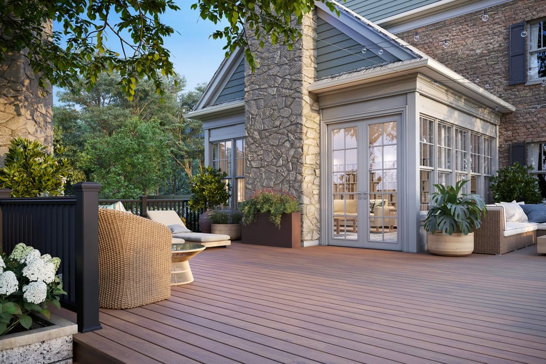 Computer-rendered reddish-brown Lineage Hatteras deck with wicker chairs beside a stone and brick house with a black railing