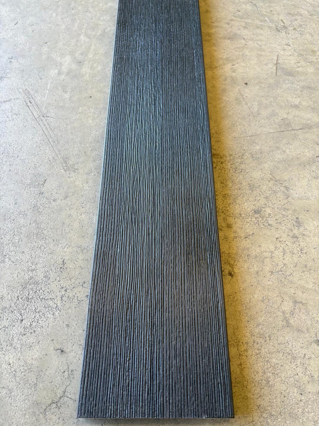 Modelo Prime Charcoal solid composite deck board lying on a concrete floor, showing its dark charcoal brushed wood-grain face