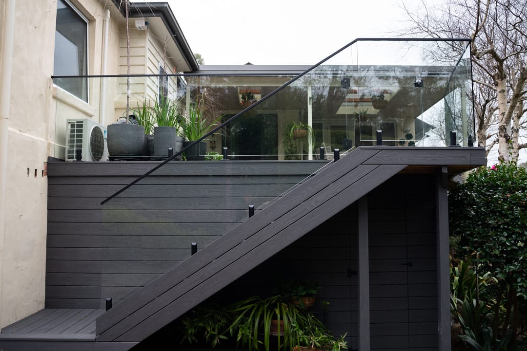Elevated charcoal composite deck and staircase with frameless glass balustrade on a rendered home, with shrubs below