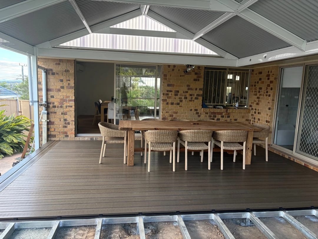 Brown composite deck under a gabled patio roof with a long timber dining table and chairs beside a brick home
