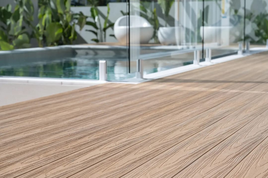 Close-up of light timber-tone COEN composite decking boards meeting a pool edge with frameless glass fencing