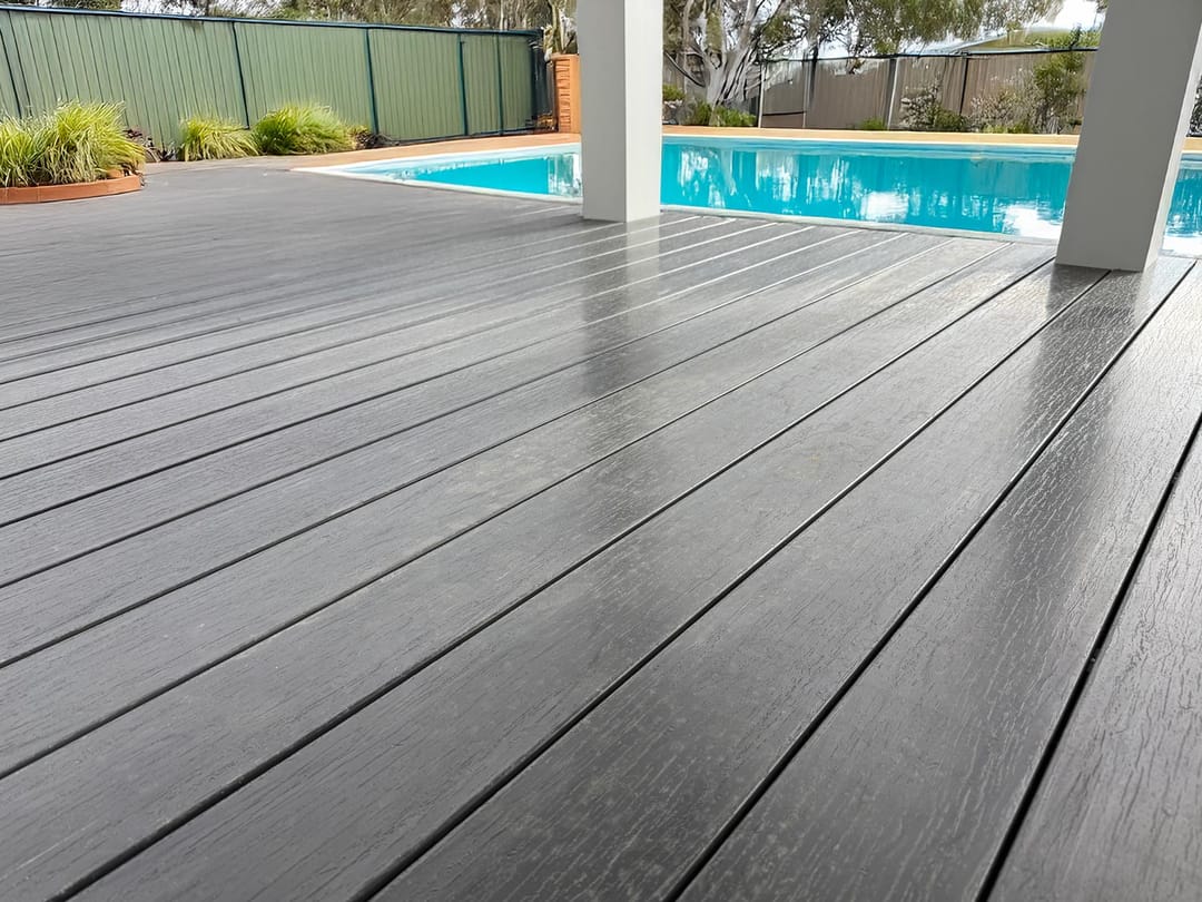 Dark charcoal OneDeck PVC deck under a verandah beside a blue pool, with garden beds behind
