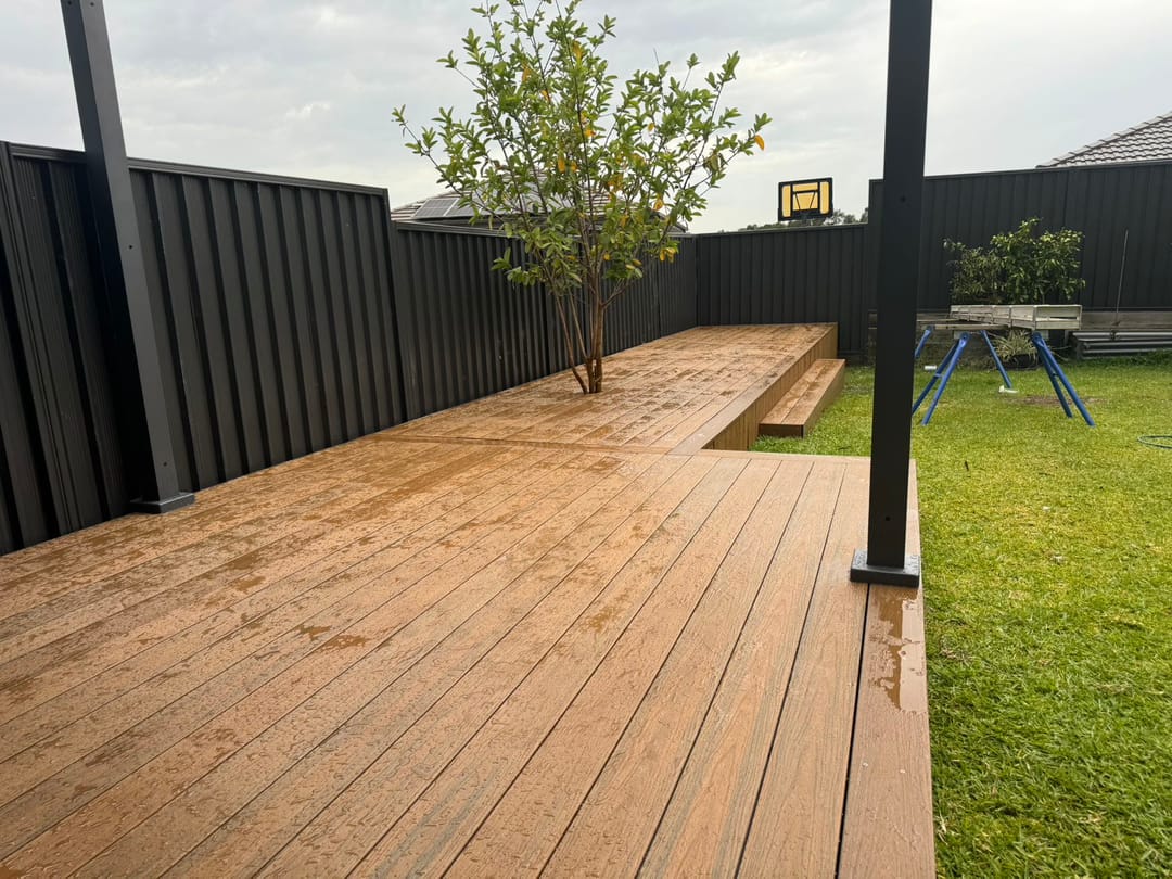 Freshly rained-on warm brown Acetech deck in a backyard, built around a young tree beside lawn and a dark steel fence