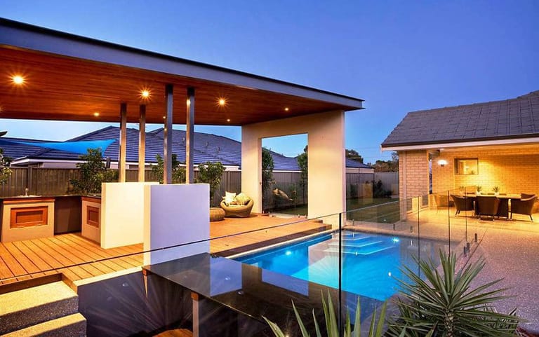 Lava Rock deck at dusk around a glass-fenced pool, with a lit alfresco kitchen and dining area behind