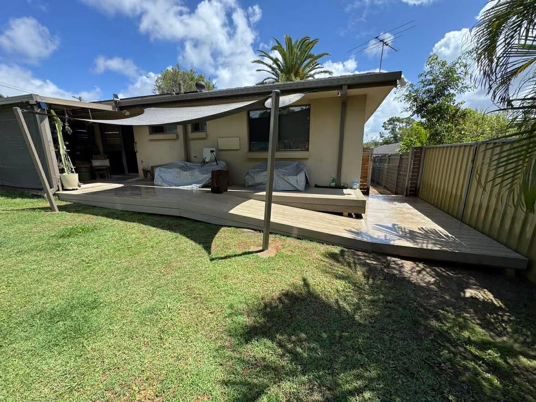 Pale greige composite deck along the back of a brick home under shade sails, with a lawn in front