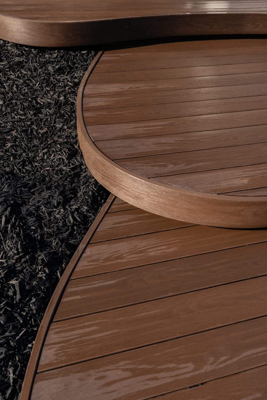 Curved WoodEvo ADVANCED composite deck edge and a round step in a warm brown tone beside black garden mulch