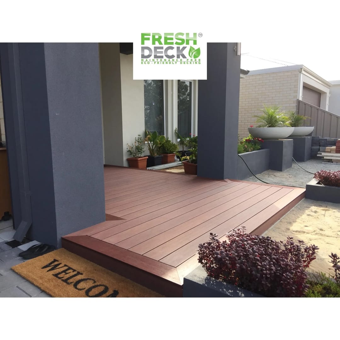 Red-brown FreshDeck composite entry deck with a picture-frame edge in front of a modern house, with a welcome mat and pot plants