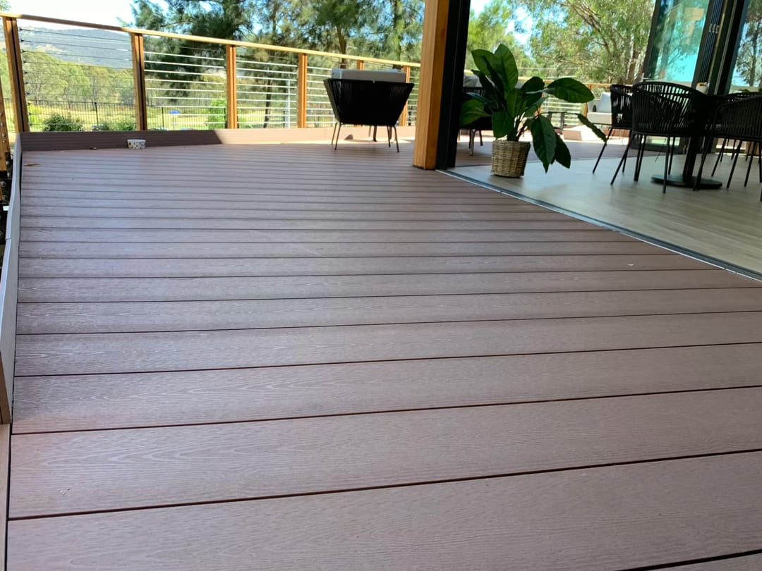 Reddish-brown wide-board composite deck on a covered terrace with cable balustrade and outdoor tables