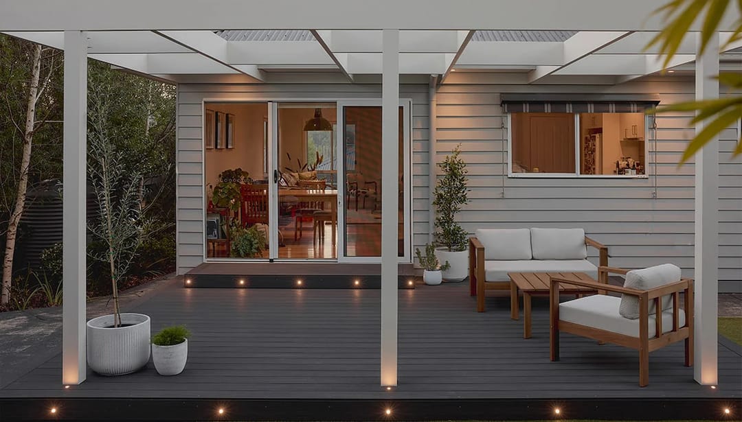 Charcoal Ekodeck Essentials Nightfall deck with lounge setting under a white pergola, lit by deck lights