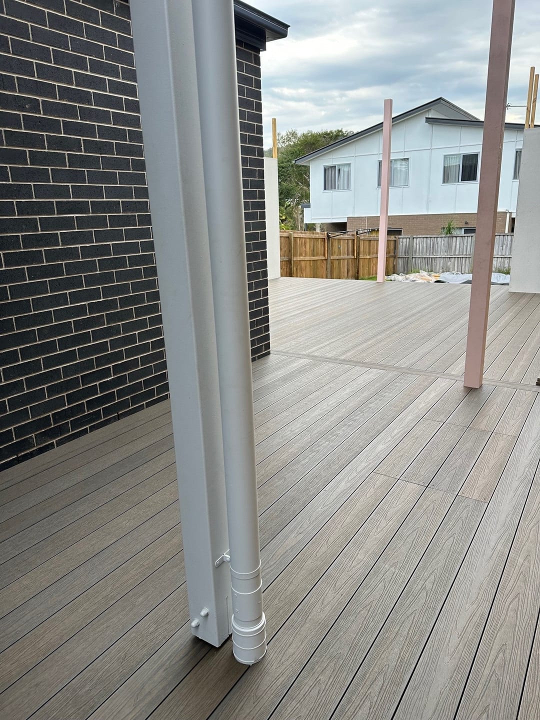 Brown composite deck beside a dark brick wall with white downpipes, looking out to neighbouring houses