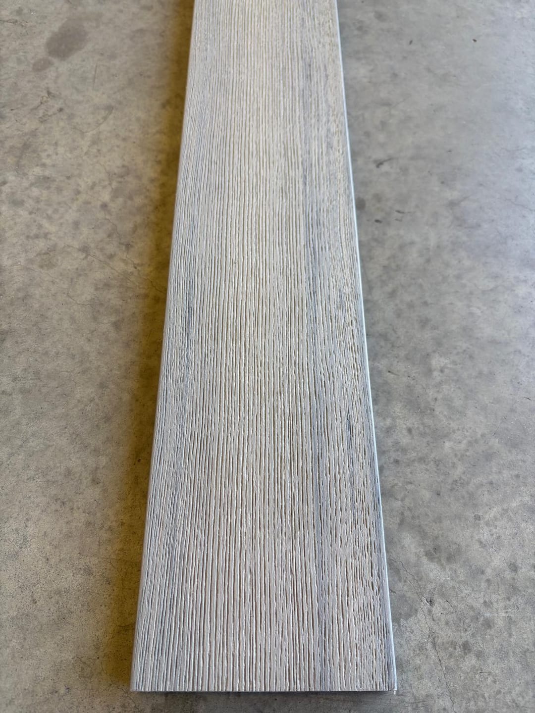 Modelo Prime Antique solid composite deck board lying on a concrete floor, showing its pale grey brushed wood-grain face